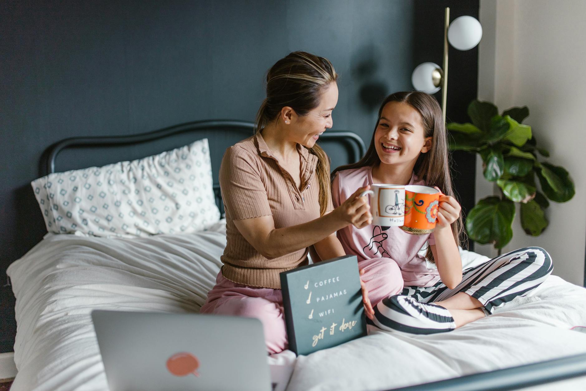 woman and a girl wearing pyjamas sitting on a bed with coffee mugs and a laptop